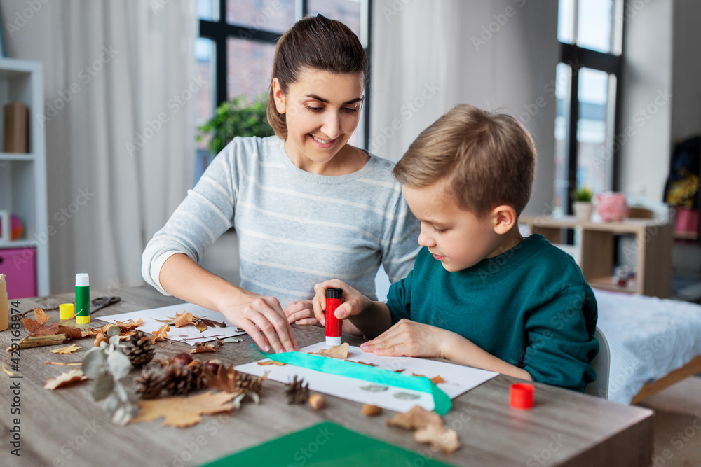 family, creativity and craft concept - mother and little son with glue sticks and paper making pictures of dry autumn leaves, pine cones and chestnuts at home