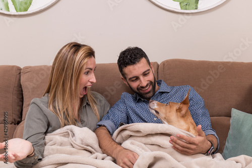 couple together with their pet, partner and their dog sitting on a sofa and covered with a blanket, the woman has an astonished face, the man smiles and the dog looks carefully at both