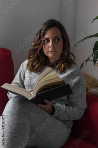 woman reading a book, young woman in gray pajamas reads a book sitting on a red sofa