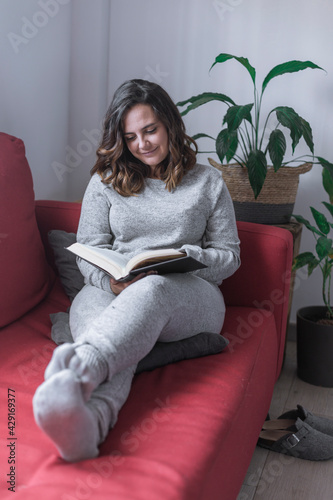 woman reading a book, young woman in gray pajamas reads a book sitting on a red sofa, there are plants around and gray slippers on the floor