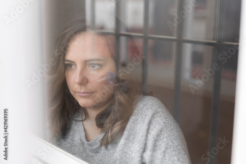 woman looking out the window, pensive woman observes the street through a closed window, the bars of the house are reflected in the glass, horizontal photo
