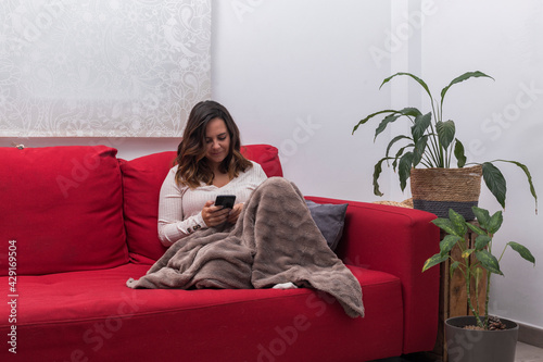 woman sends messages with the mobile, brunette woman sitting on a red sofa and covered with a blanket writes with the mobile and smiles, next to the sofa there are plants, horizontal photo