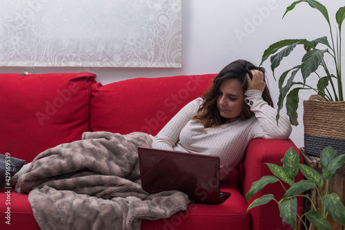woman works with laptop at home, brunette woman sitting on a red sofa and covered with a blanket writes on the computer, next to the sofa there are plants, horizontal al photo