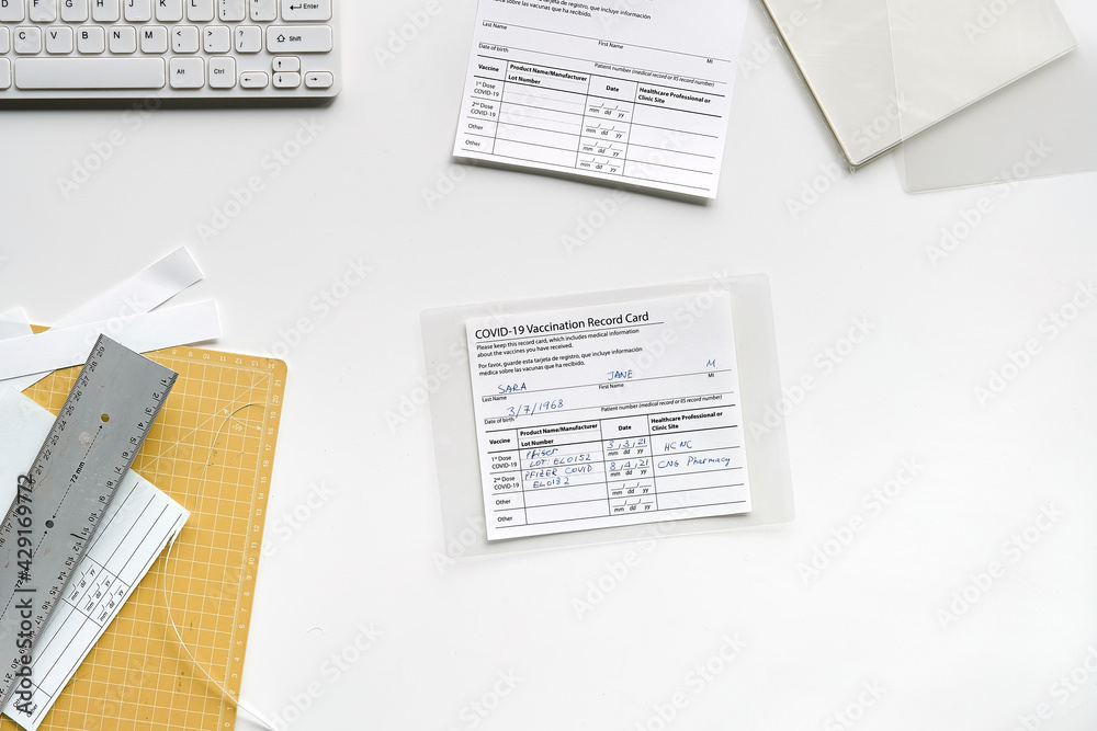 Obraz premium Fake covid vaccination record card. Covid scams. Forged health certificate with false vaccination record against covid-19. Blank card, pen, paper knife,laminator. Top view, flat lay on white table.