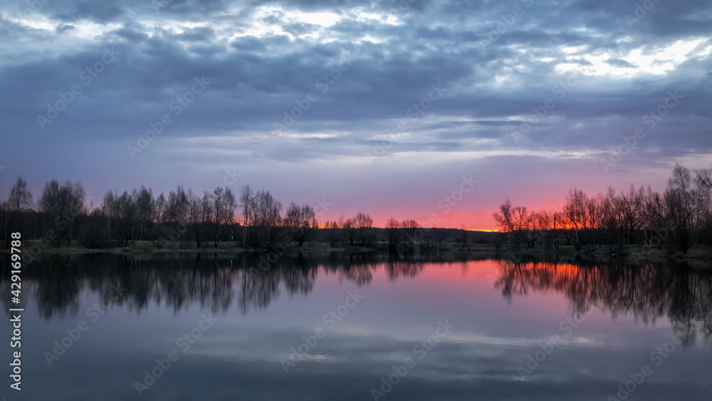 Dramatic pink sunset over forest lake with bare tree silhouettes on horizon reflected on water surface