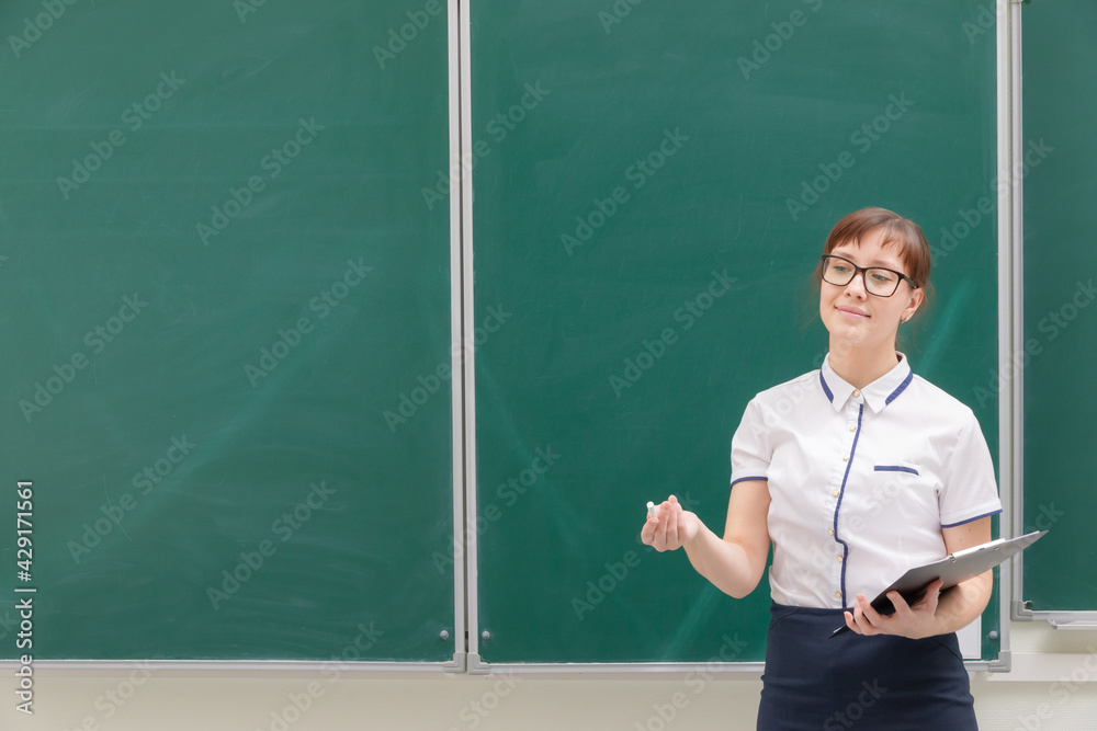 school teacher young pretty woman in a white blouse with a folder and chalk in her hands in the classroom on the background of the blackboard. portrait