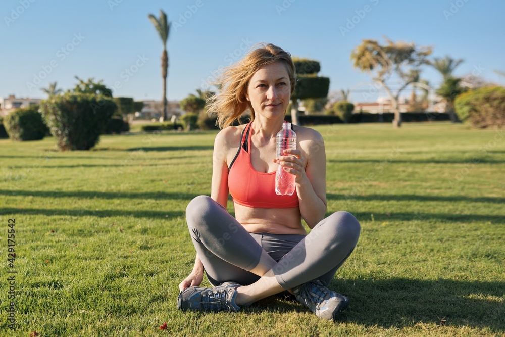 Mature woman doing morning exercises on green grass in park, with bottle of water Mature woman doing morning exercises on green grass in park, with bottle of water