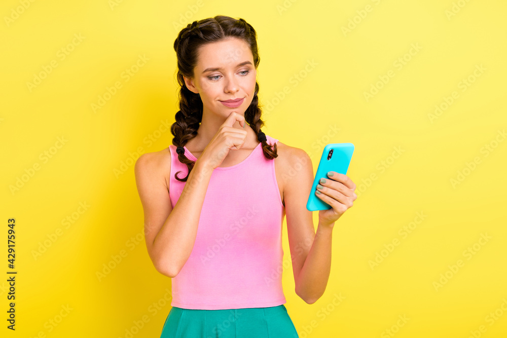 Photo of cute thoughtful young woman pigtails wear tank top finger chin looking modern device isolated yellow color background Photo of cute thoughtful young woman pigtails wear tank top finger chin looking modern device isolated yellow color background