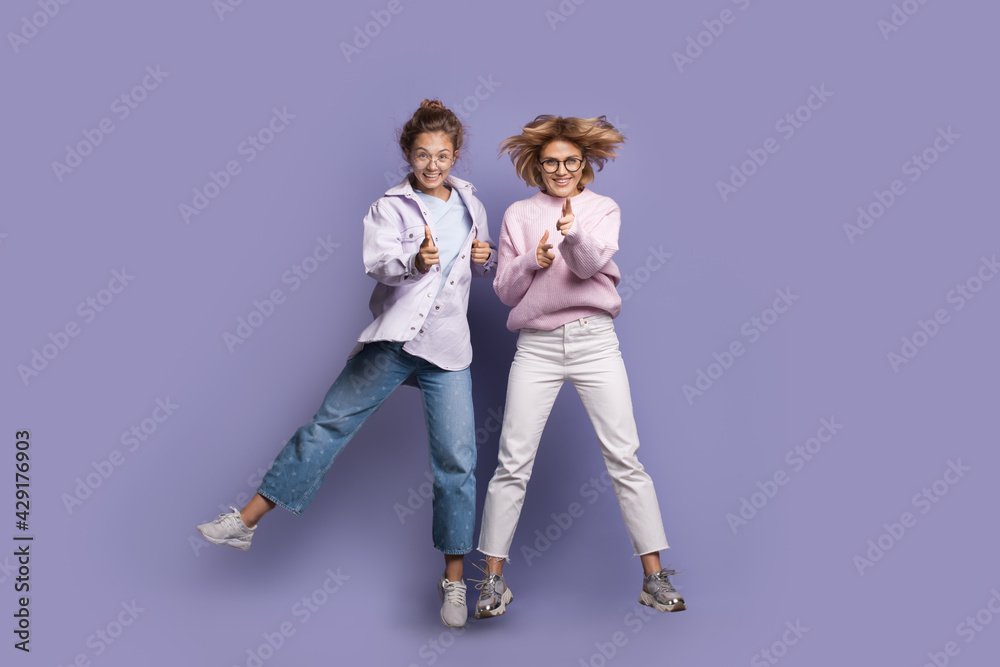 Fototapeta premium Blonde sister are jumping on a purple wall smiling and gesturing the gun sign