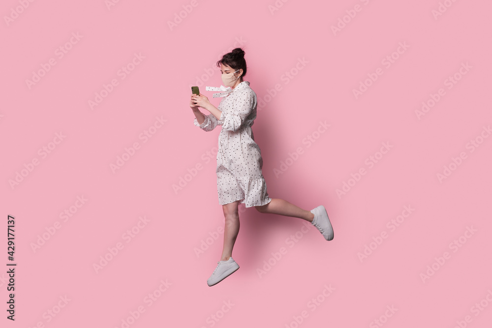 Fashionable woman with medical mask is posing on pink studio wall chatting on mobile