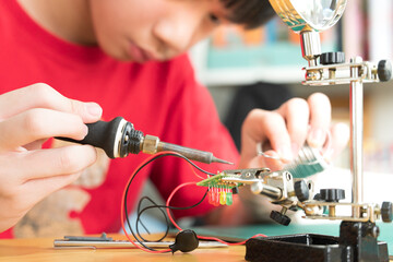 STEM education and 21st century skills concept. Young Asian teenager, kid is soldering water moisture sensor circuit board with grip hand desktop stand. DIY school science and technology project.