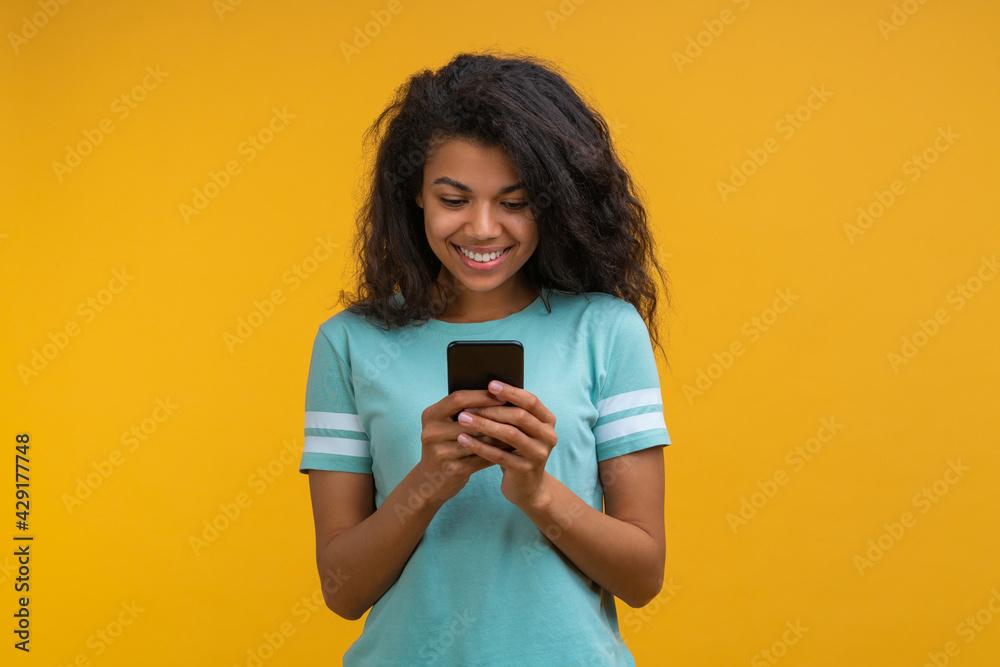 Half-length studio portrait of beautiful casually dressed dark skinned girl with perfect charming smile texting message via her mobile phone isolated on bright colored yellow background Half-length studio portrait of beautiful casually dressed dark skinned girl with perfect charming smile texting message via her mobile phone isolated on bright colored yellow background
