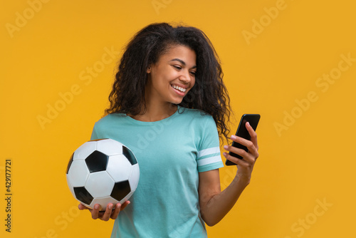 Beautiful smiling dark skinned girl holding soccer ball and watching online broadcast on his mobile phone cheering for favourite team, making bets online and waiting for winning results photo