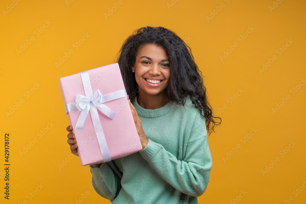 Portrait of happy young african american girl holding gift box decorated with satin ribbon with broad sincere smile Portrait of happy young african american girl holding gift box decorated with satin ribbon with broad sincere smile
