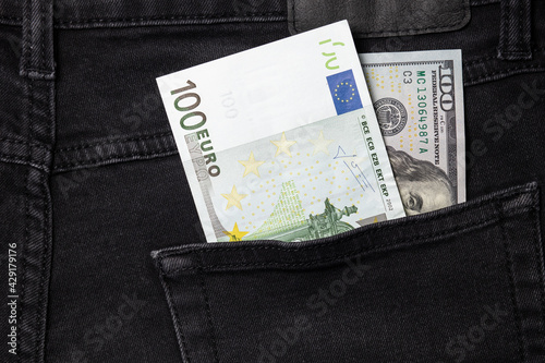  One hundred euro and one hundred dollar bills sticking out of the pocket of black jeans
