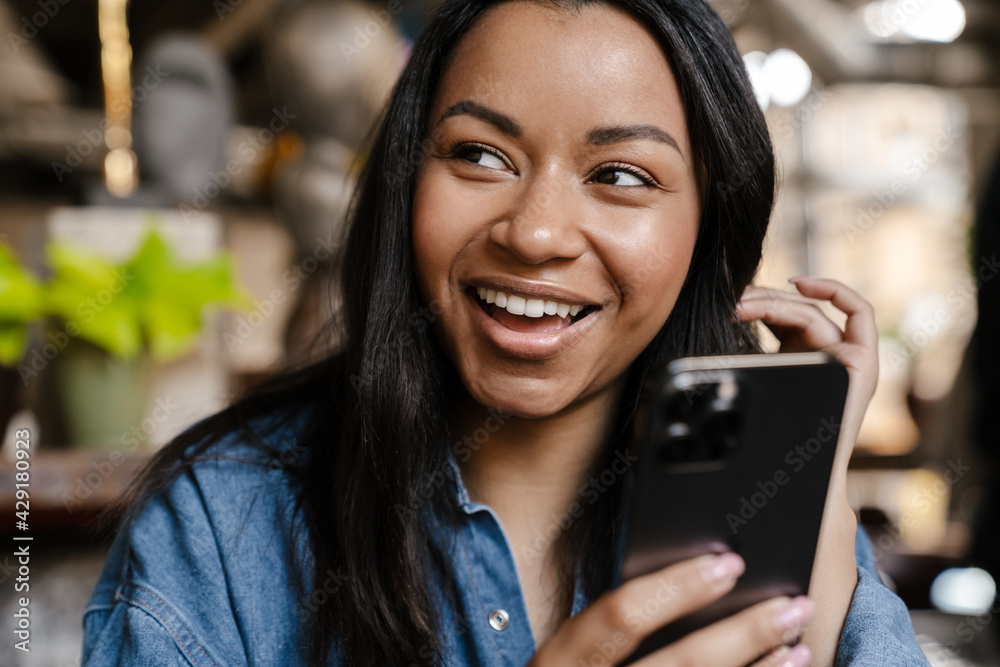 Black young woman smiling and using cellphone while sitting in cafe