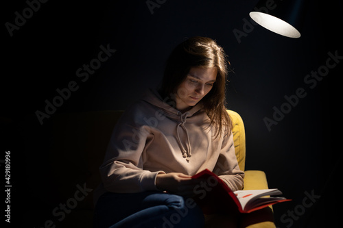Attractive long-haired girl intently reads book sitting knees bent under spot lamp. pretty lady reading with concentration love story historic novel. female reader reading copy space on black bacdrop photo