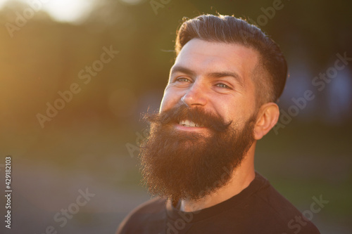 Laughing brutal stylish adult man with a long black beard and mustache in a dark shirt against the sunset sky