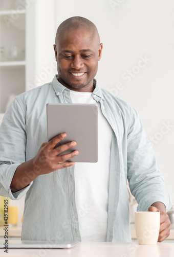 Leisure time. Happy black man reading e-book on digital tablet and drinking coffee in kitchen