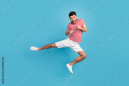 Full length photo of cheerful strong young guy wear red outfit jumping high practicing karate isolated blue color background