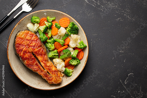 Healthy fish dinner: grilled salmon fish steak with vegetables salad on ceramic plate with fork and knife on black slate stone background from above with copy space, clean and diet eating 