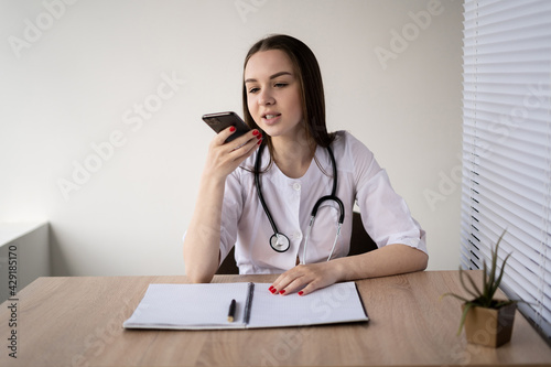 Photos Caucasian lady doctor sitting at her working table communicating with a virtual assistant on a modern smartphone