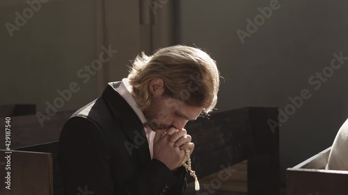 Lockdown portrait of young Caucasian ginger man wearing black suit sitting on wooden pew in Christian church, holding rosary beads and praying to God