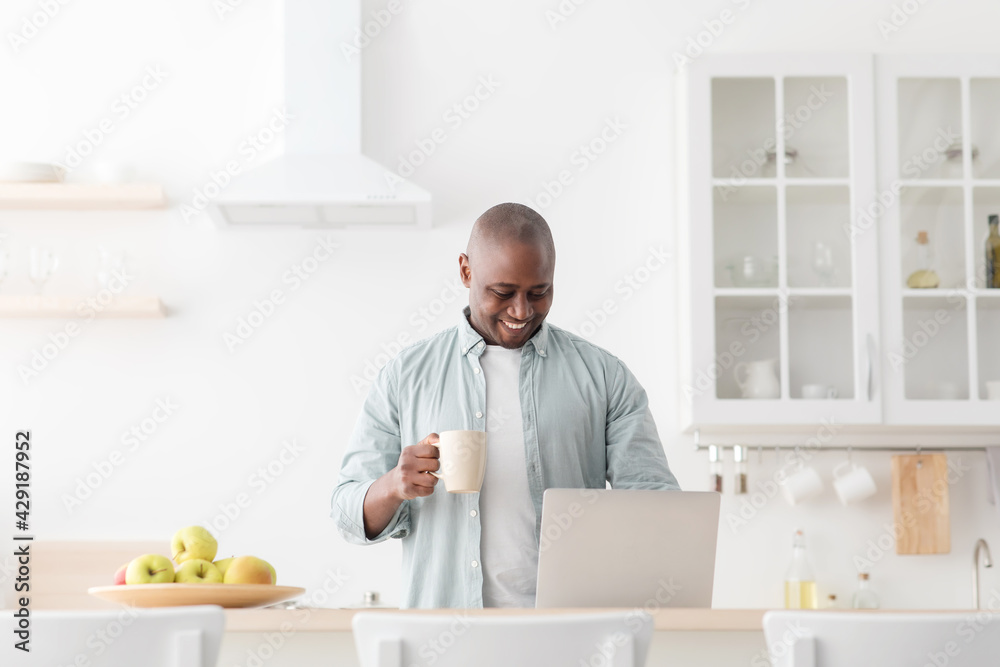 Excited mature black man using laptop in kitchen and drinking coffee, browsing social media or reading online news Excited mature black man using laptop in kitchen and drinking coffee, browsing social media or reading online news