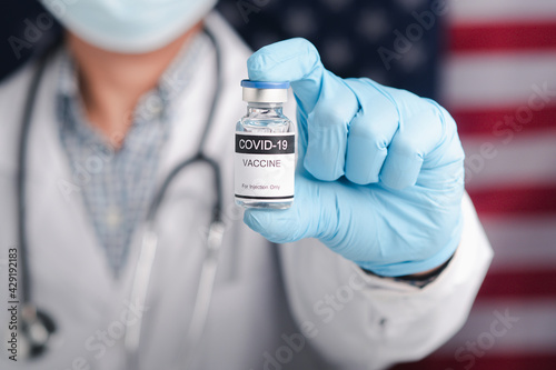A doctor holding a COVID-19 vaccine bottle against the background of the United States flag. Vaccine for immunization and treatment from virus infection.