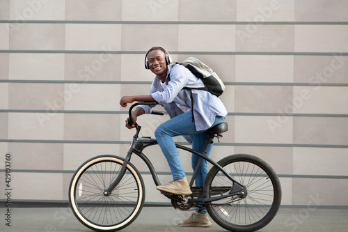 Joyful black guy with backpack and headphones riding bicycle near brick wall on city street