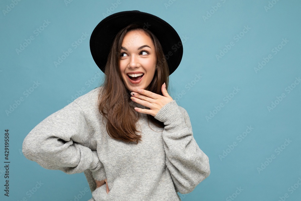 Portrait of young positive happy surprised beautiful brunette woman with sincere emotions wearing stylish grey pullover and black hat isolated over blue background with copy space Portrait of young positive happy surprised beautiful brunette woman with sincere emotions wearing stylish grey pullover and black hat isolated over blue background with copy space