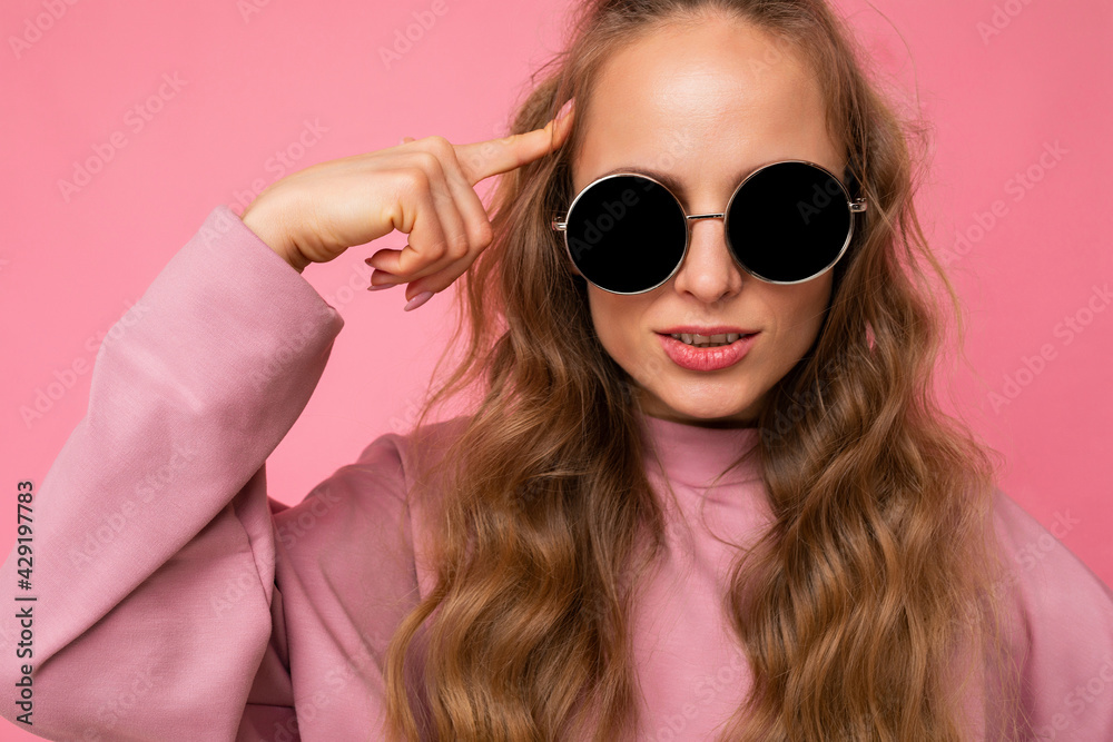 Closeup shot of beautiful serious young dark blonde curly woman isolated over pink background wall wearing casual pink sport clothes and stylish sunglasses looking at camera Closeup shot of beautiful serious young dark blonde curly woman isolated over pink background wall wearing casual pink sport clothes and stylish sunglasses looking at camera
