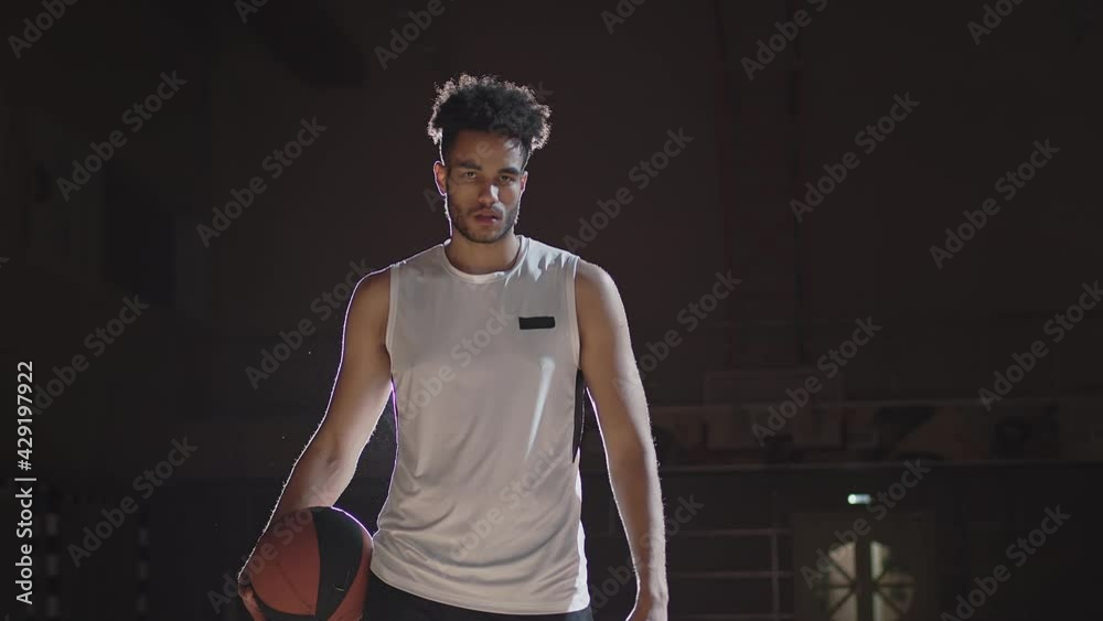Portrait slowmo tracking shot of young African-American player with basketball posing for camera on dark indoor court with spotlight shining behind him Portrait slowmo tracking shot of young African-American player with basketball posing for camera on dark indoor court with spotlight shining behind him