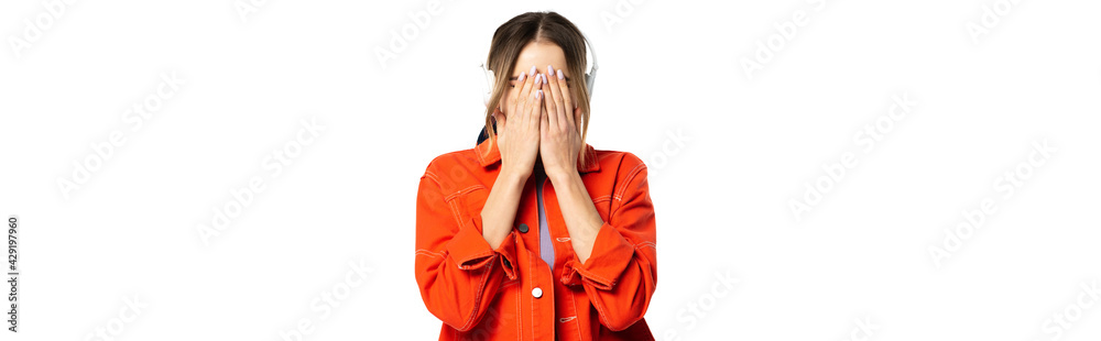 woman in orange shirt covering face while hands while listening music in wireless headphones isolated on white, banner