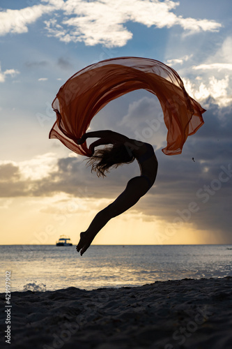 Slika na platnu Flexible fit woman jumping with silk during dramatic sunset with stormy clouds