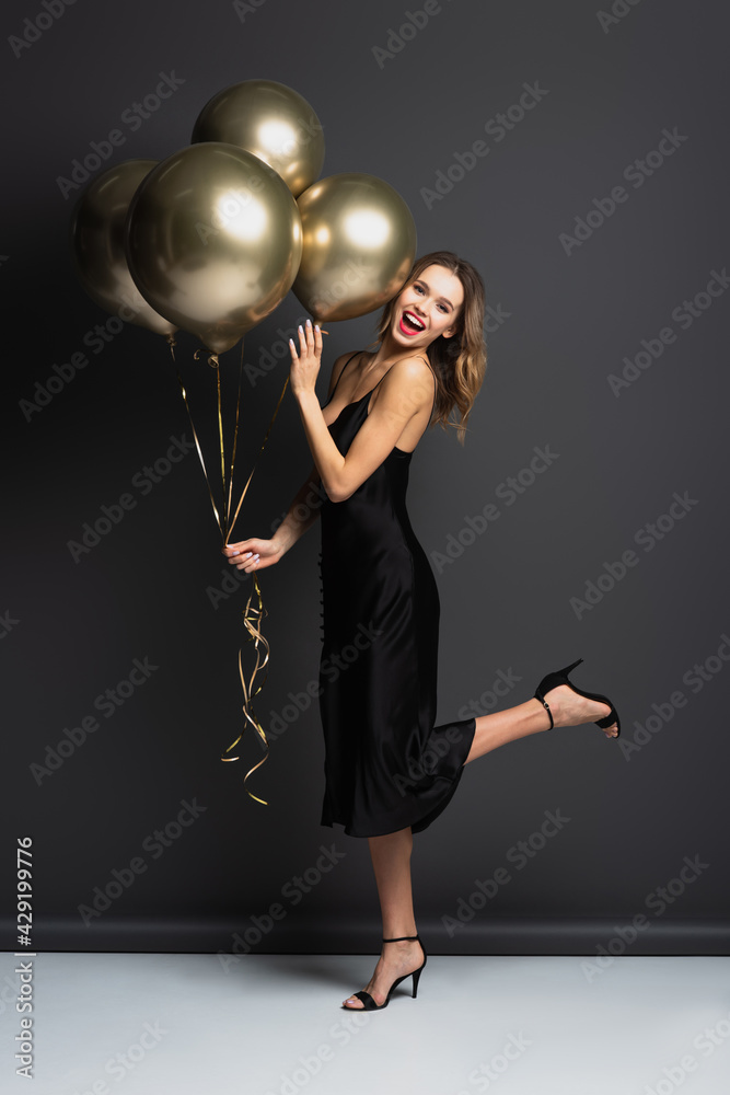 full length of amazed young woman in black slip dress holding golden balloons on grey full length of amazed young woman in black slip dress holding golden balloons on grey