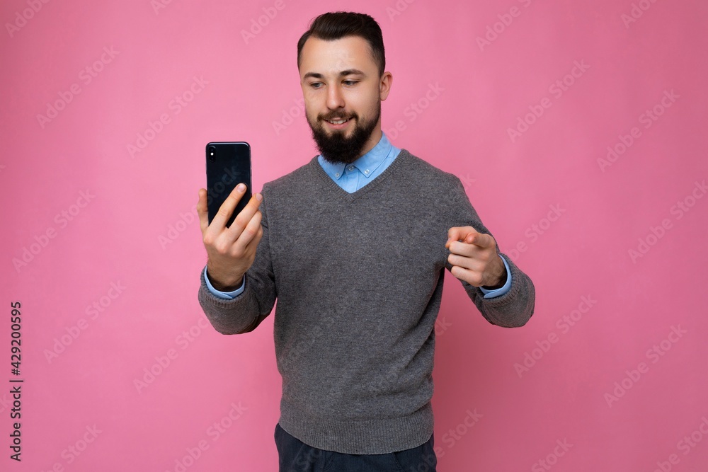 Handsome happy cool young brunette unshaven man with beardwearing stylish grey sweater and blue shirt standing isolated over pink background wall holding smartphone and using phone looking at gadjet Handsome happy cool young brunette unshaven man with beardwearing stylish grey sweater and blue shirt standing isolated over pink background wall holding smartphone and using phone looking at gadjet
