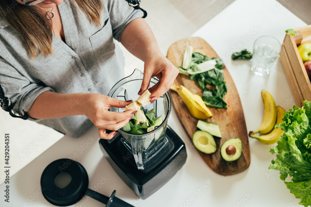 Woman is preparing a healthy detox drink in a blender - a  green smoothie with fresh fruits, green spinach and avocado. Healthy eating concept, ingredients for smoothies on the table, top view - obrazy, fototapety, plakaty 