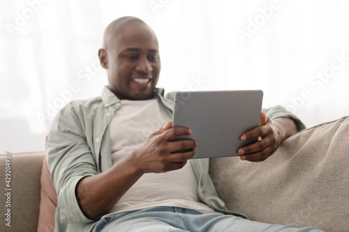 Positive african american mature man spending time with digital tablet, browsing internet while resting on comfy couch