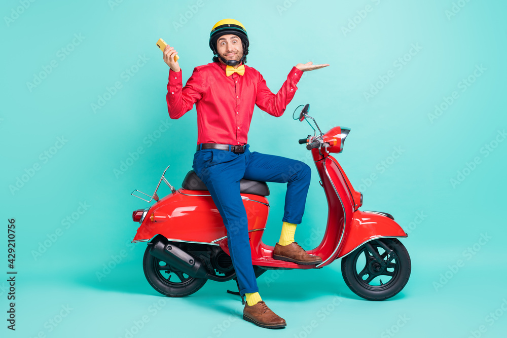 Full size photo of young hesitant man on scooter hold phone shrug shoulders doubtful isolated on teal color background Full size photo of young hesitant man on scooter hold phone shrug shoulders doubtful isolated on teal color background