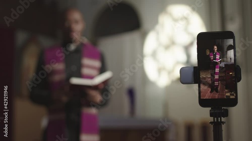 Racked focus shot of young African-American pastor wearing robe and white collar standing in Christian church in front of smartphone with Holy Bible in hands and preaching sermon