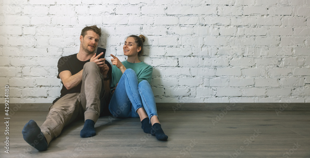 couple sitting on floor by the wall at empty room and using phone to choose furniture online. moving to new home concept