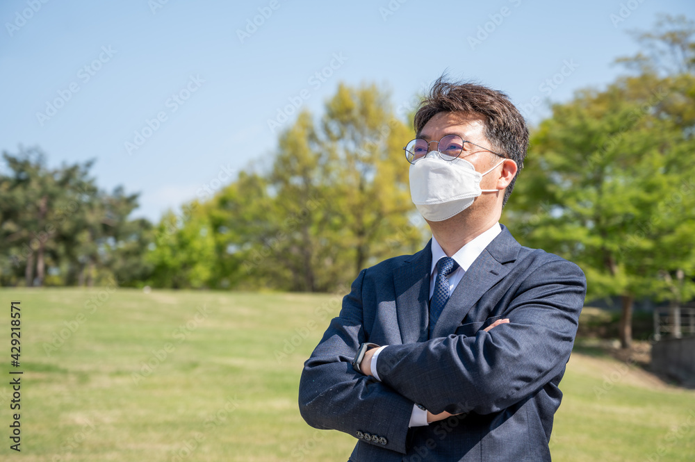 Asian middle-aged businessman with folded arms wearing a mask on the lawn. Asian middle-aged businessman with folded arms wearing a mask on the lawn.
