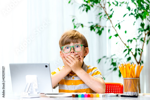 Blond kid with freckles in green eyes. Covering mouth with hands, shocked and afraid for mistake. surprised expression on kids face. Big eyes. Online school lesson. Remote education on laptop
