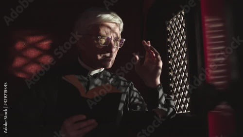 Medium shot of old Caucasian priest wearing black robe, white collar and eyeglasses sitting in confessional with Holy Bible in hands and listening to confession photo