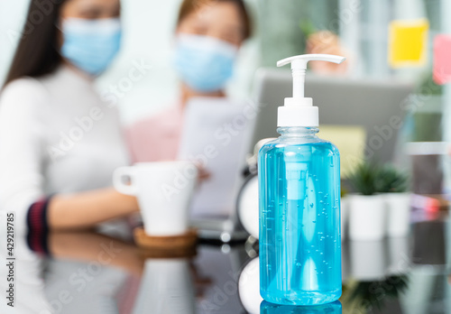 A bottle of alcohol gel is set on an office desk with a blur female employee wearing a surgical mask background. Alcohol gel for washing hands during the outbreak of the virus in health care concept.