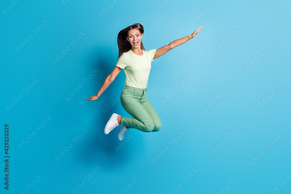 Full length photo of charming sweet young woman wear green outfit jumping arms sides isolated blue color background