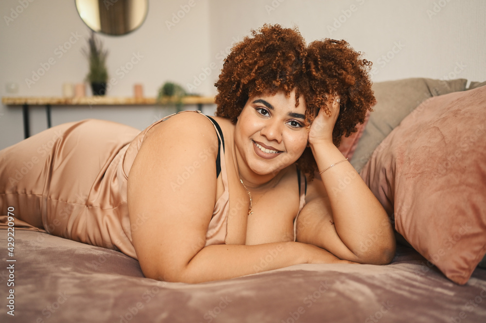 Naklejka premium Beautiful curvy plus size African black woman afro hair lying on bed in silk powder pink dress cozy bedroom interior design. Body imperfection, body acceptance, body positive and diversity concept.