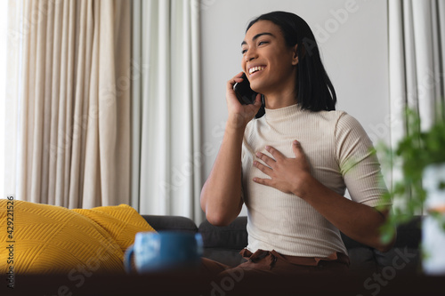 Happy mixed race transgender woman relaxing in living room sitting on couch talking on the phone