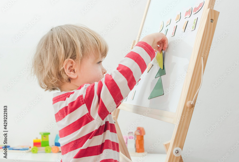 Cute little child playing at a magnetic board, learning letters, numbers and geometric shapes at home. Educative and funny indoor activity for early development.